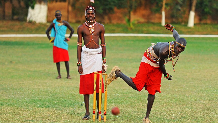 Maasai Warrior bowler sends down a delivery as Maasai umpire looks on.