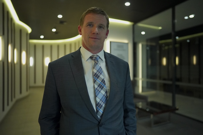 A man in a suit and tie stands in the foyer of an office building, looking into the camera.