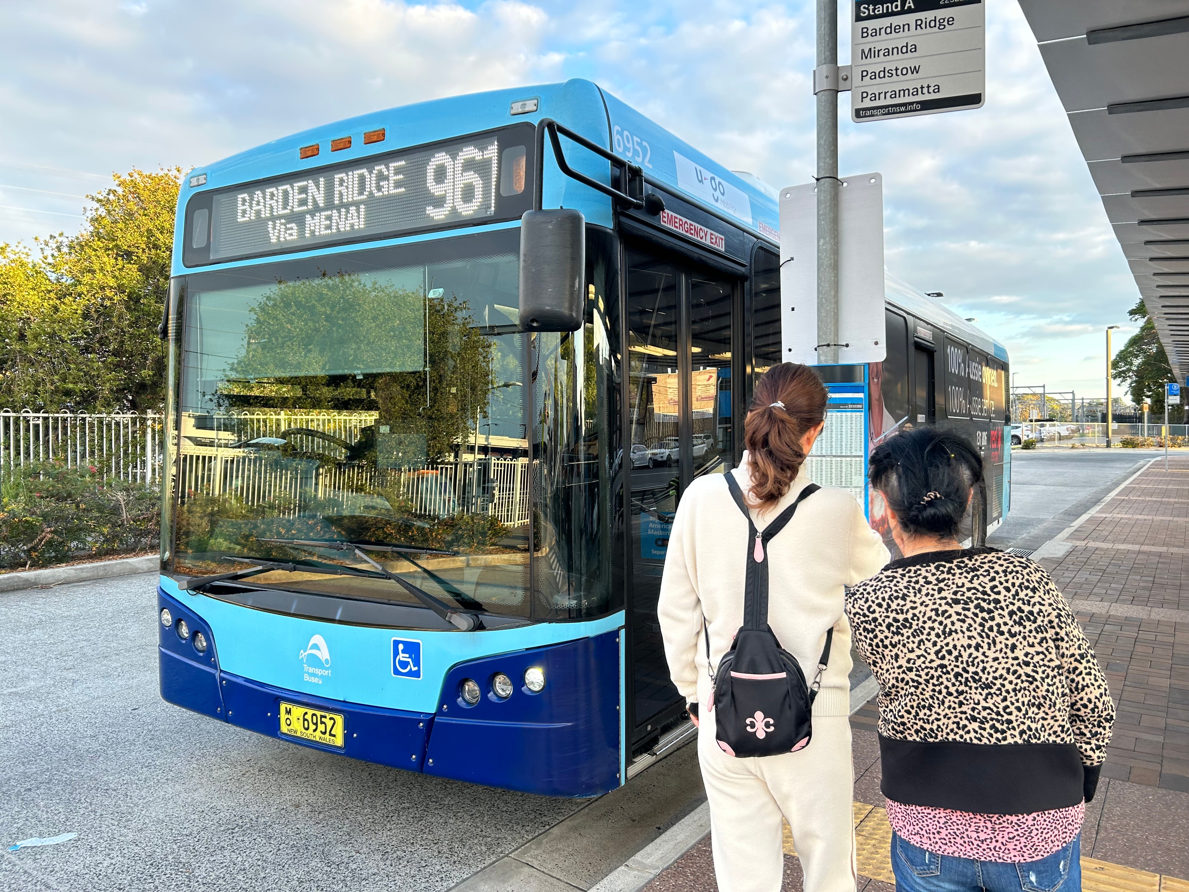 A blue bus at a bus stop with two people looking at a timetable.