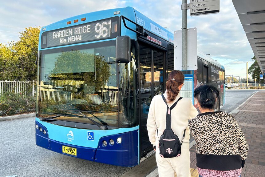 A blue bus at a bus stop with two people looking at a timetable.