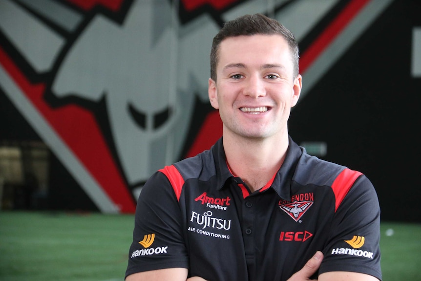 Conor McKenna stands arms crossed smiling at the camera.