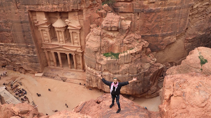 A man stands on sandstone, with a carving of a temple in the background, travelling through Petra, Jordan on his own.