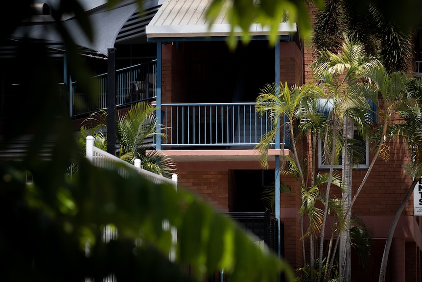 The exterior of a two-storey red brick building with blue balcony railings, seen through trees. 