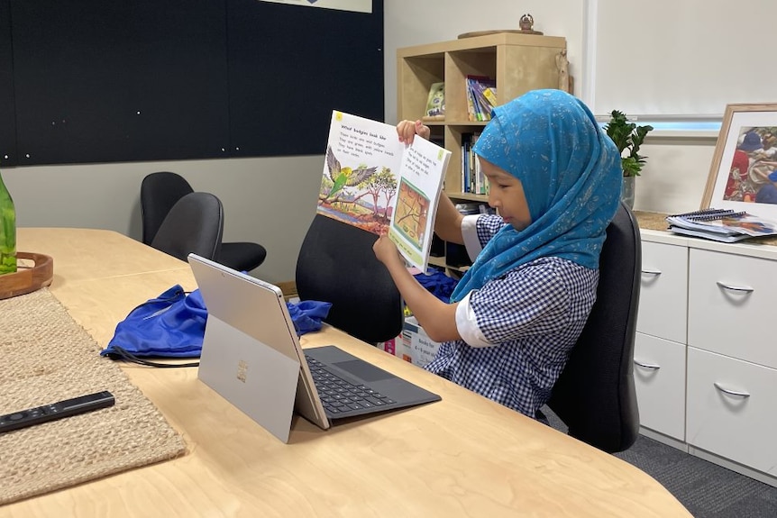 A girl holds up a book at a computer.