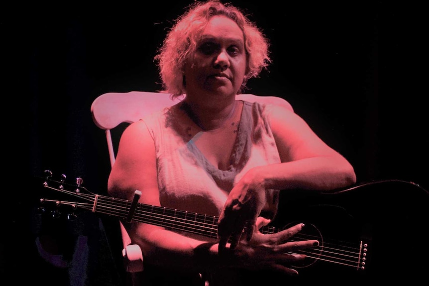 Woman sitting in chair on stage with guitar looking at camera, dark pink mood lighting