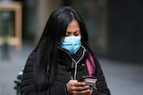 A woman wearing a mask and a winter coat looks down at her phone.