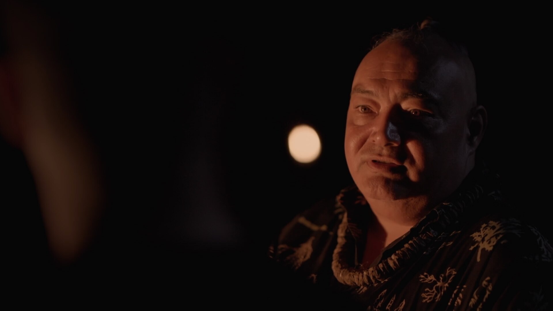 Akoni sits in the darkness outside with his face half bathed in light with a serious expression.