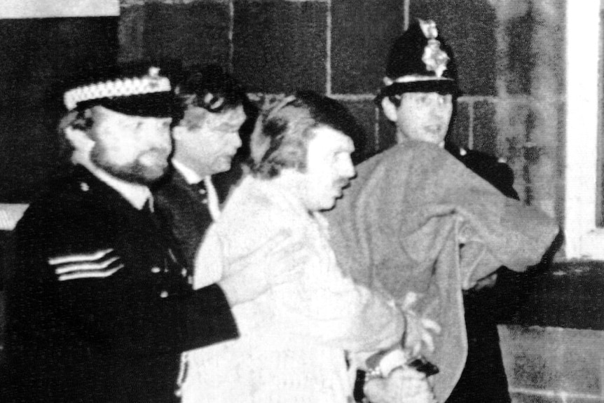 A black and white photo of the Yorkshire Ripper, with a blanket over his head, being taken into custody.