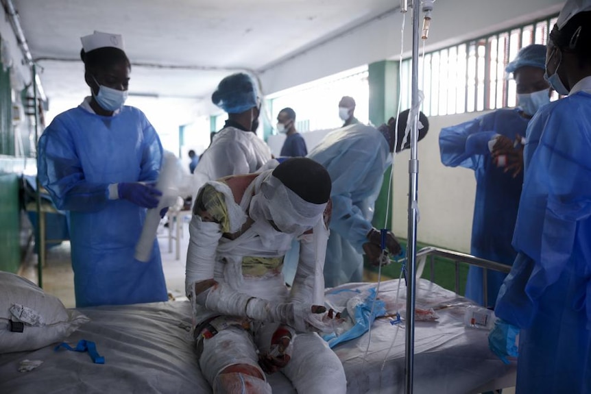 Un bărbat învelit în tifon și bandaje stă cu capul în jos pe un pat de spital, în timp ce medicii lucrează în jurul lui 
