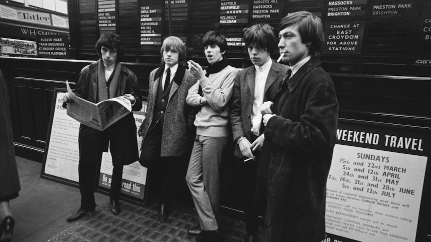 The Rolling Stones stand in front of a departures board in a train station, London, 1964