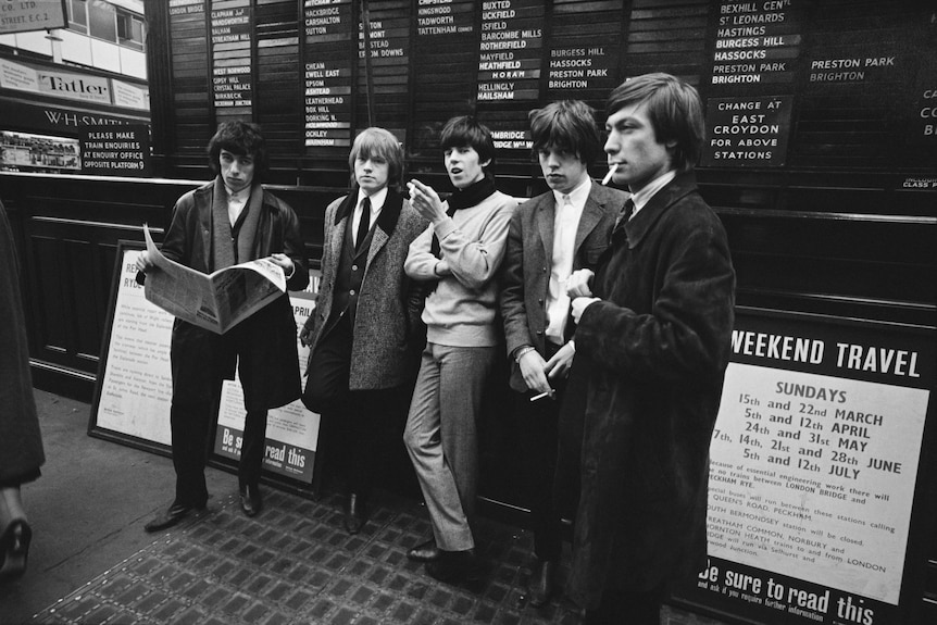 The Rolling Stones stand in front of a departures board in a train station, London, 1964