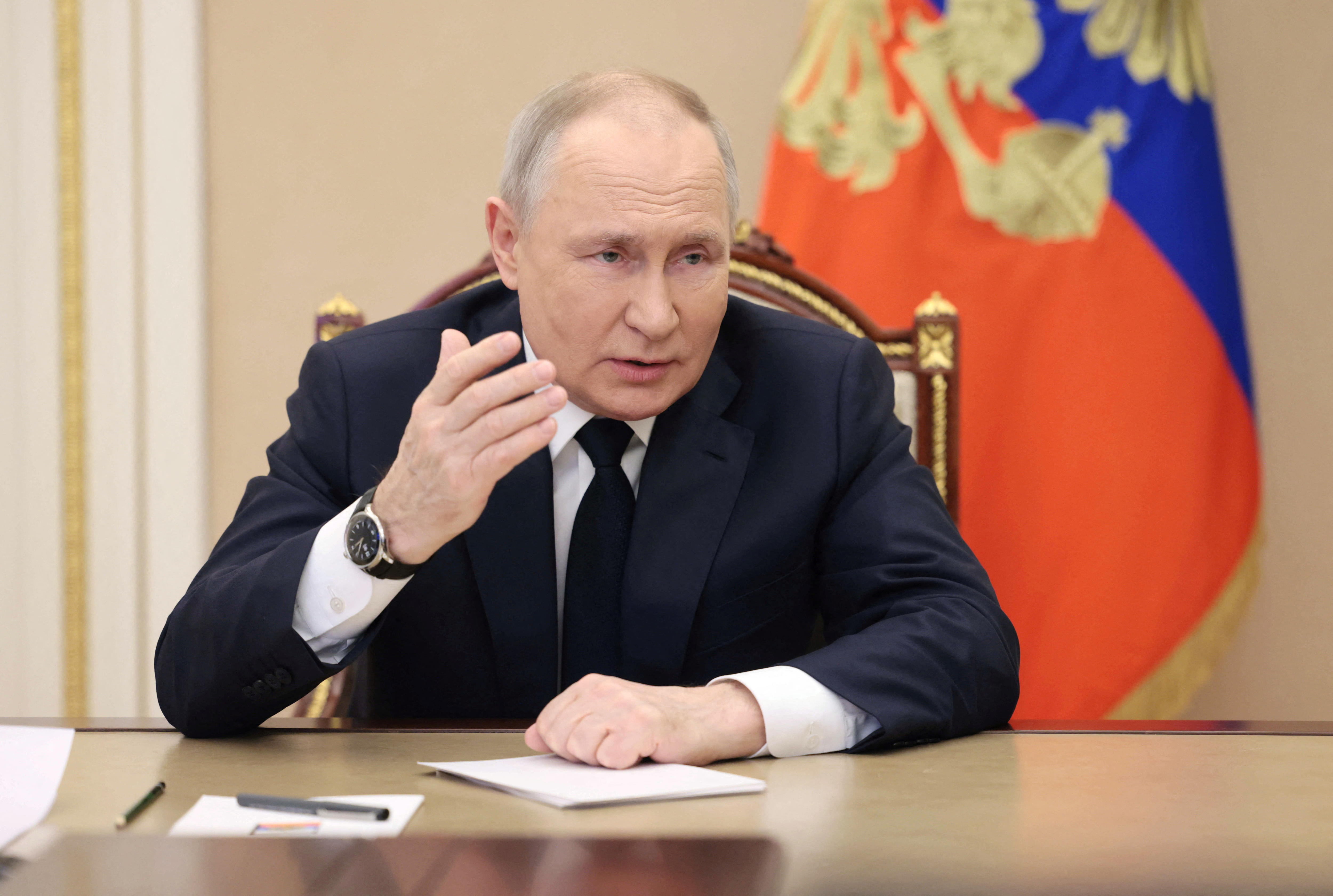 An older white man with thinning hair in a suit speaks while seated in front of a gold-trimmed blue and red flag.