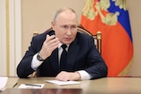 An older white man with thinning hair in a suit speaks while seated in front of a gold-trimmed blue and red flag.