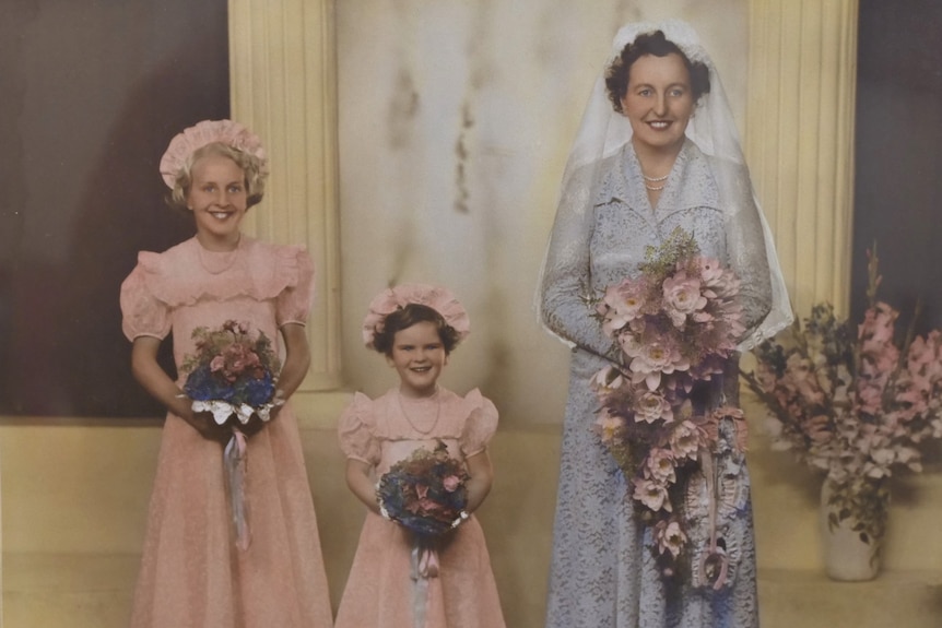A bride in a collared blue lace dress with a light blue veil stands with two flower girls dressed in pink. 