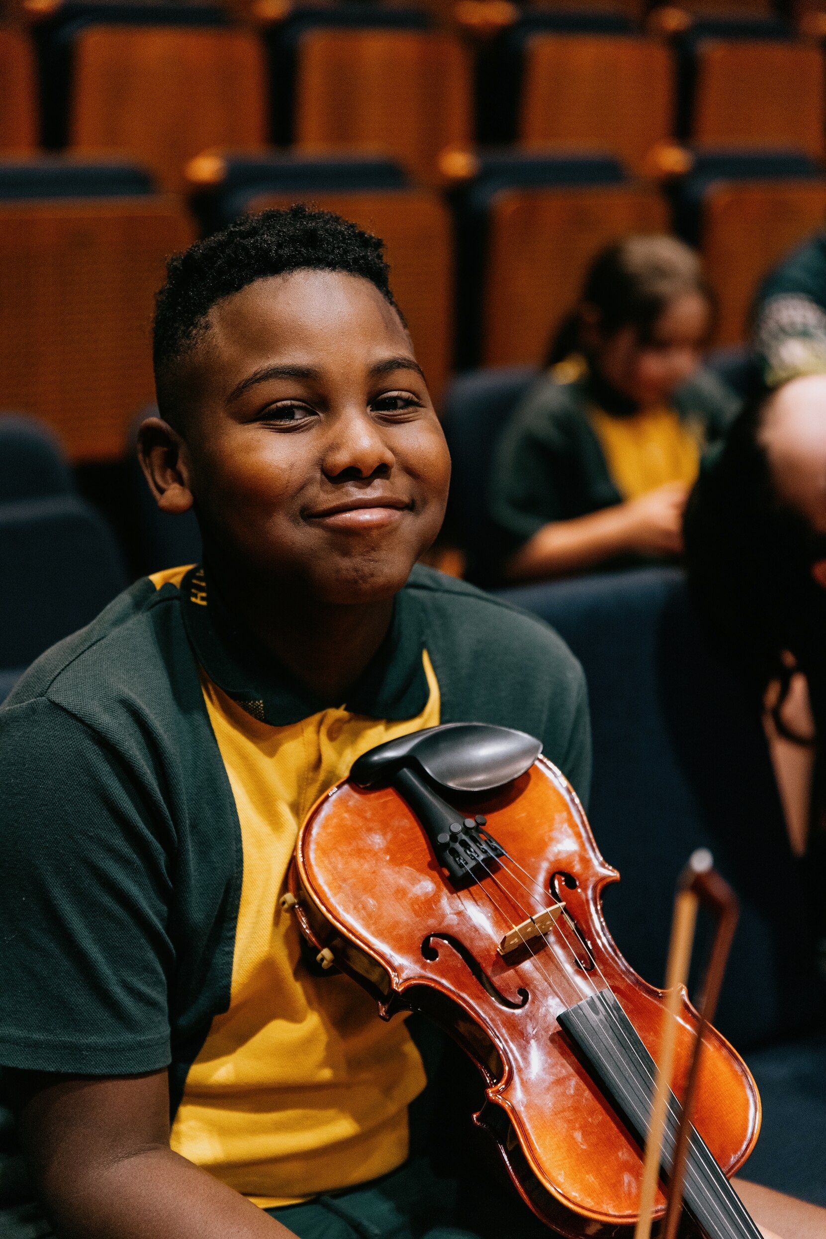 A primary-aged Black boy in a yellow and green school uniform smiles sweetly while holding a violin in an auditorium.
