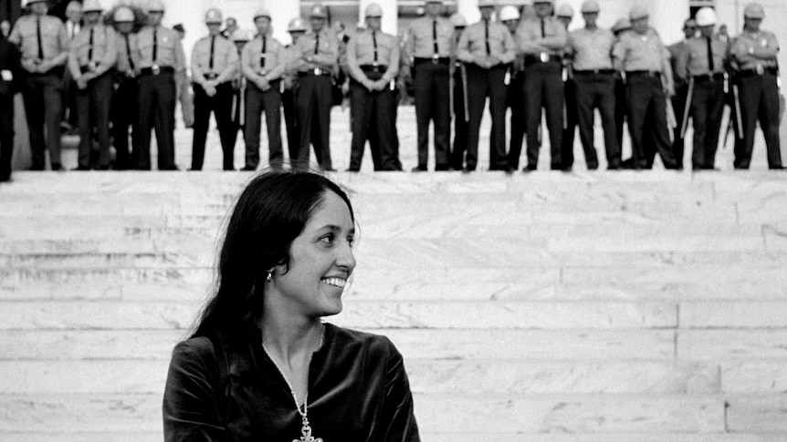 Joan Baez smiles at the bottom of the stairs at a government building, a large line of police stands at the top.
