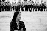 Joan Baez smiles at the bottom of the stairs at a government building, a large line of police stands at the top.