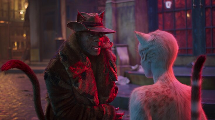 A dark brown CGI cat with man's face wears fur coat and hat, pleads with cream CGI cat standing in pink neon lit cobbled street.