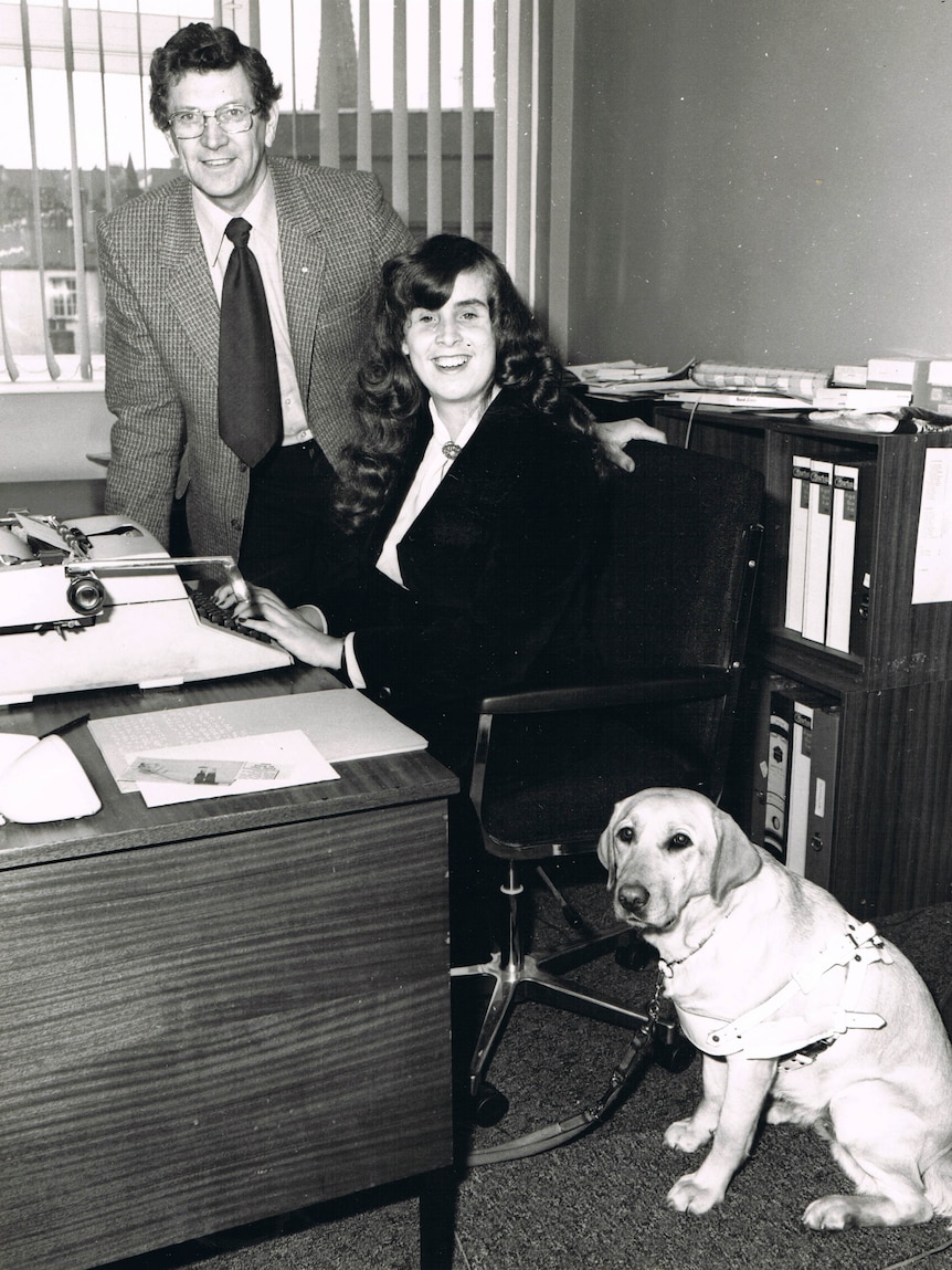 Black and white photo of Elaine. She is sitting at a desk wearing a dark suit. Her hair is very long. Her guide dog sits nearby.