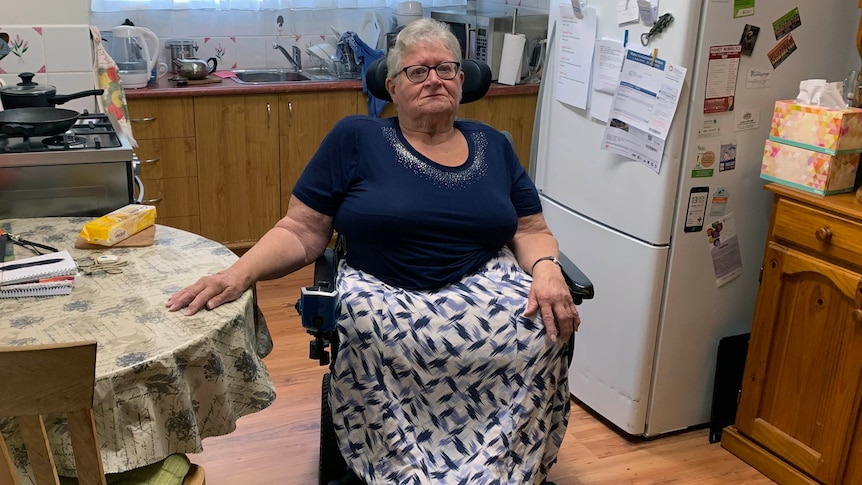 Lyn Bates sitting in a wheelchair in her kitchen with her hand on the table.