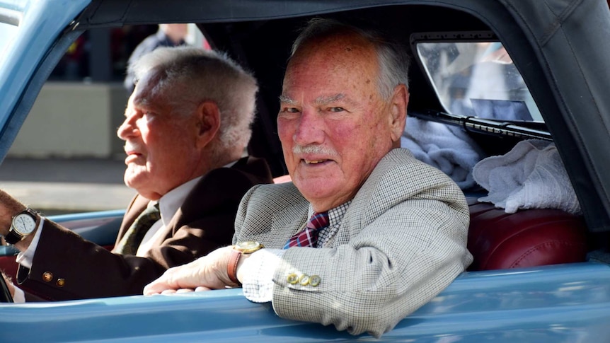 An old man in a grey knit suit and red tie rests his elbow from the passenger seat of an old blue car, smiling.