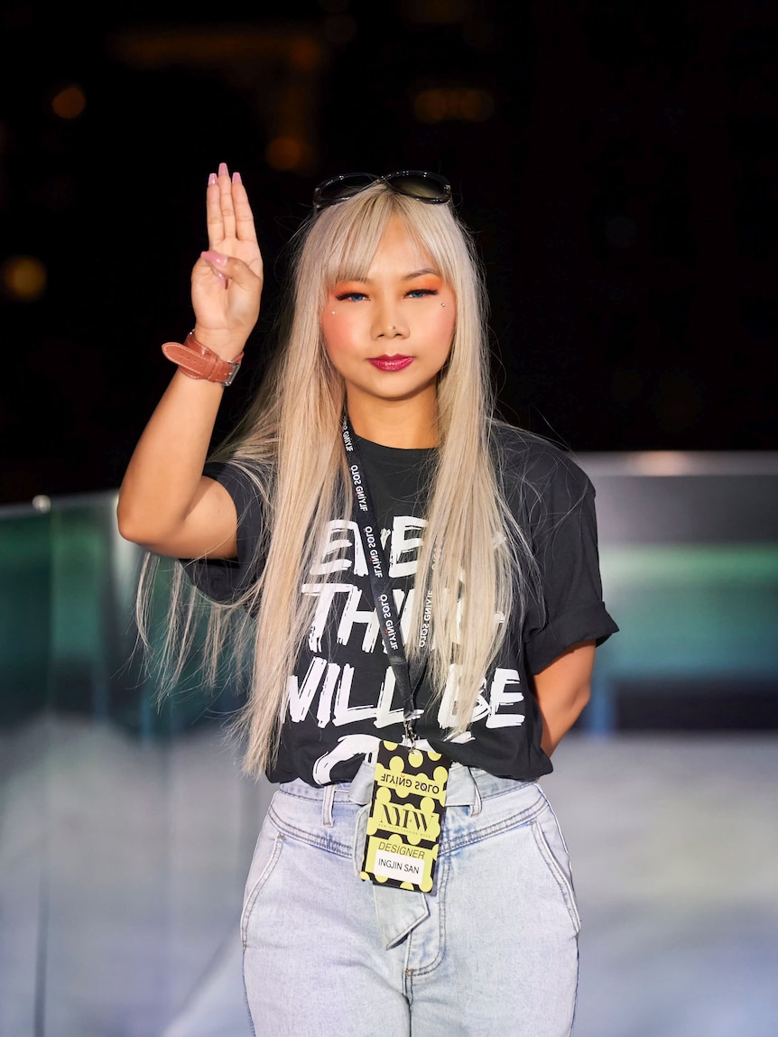 A woman with long blonde hair wearing a black t-shirt and holding up a three-finger salute