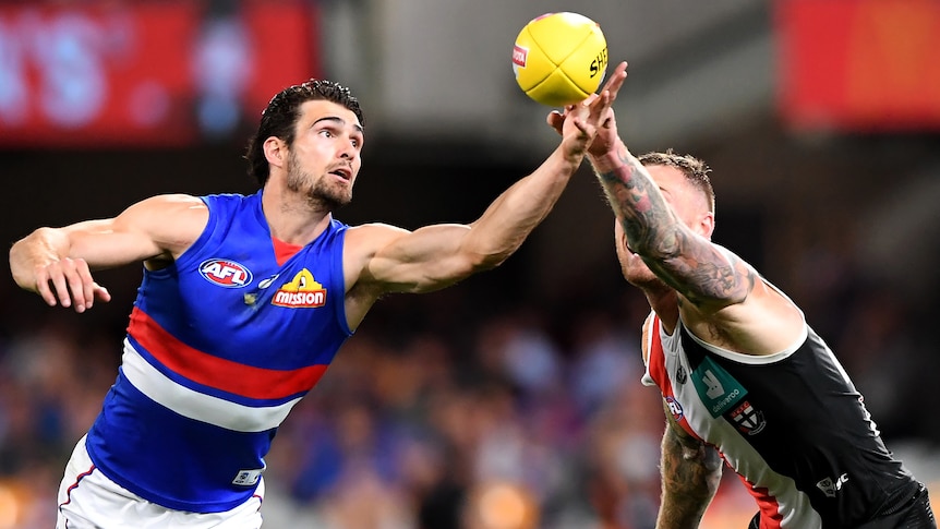 A man in a blue AFL jersey reaches out for a yellow ball, in front of a man in a red, black and white jersey.