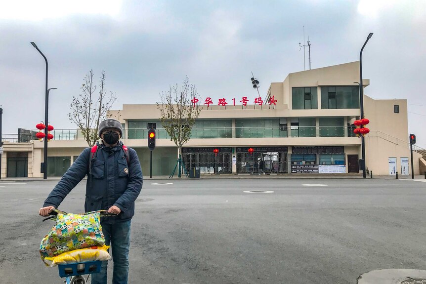 A man in a black face mask stands with a bicycle in front of an empty street and building with hanging red lanterns.