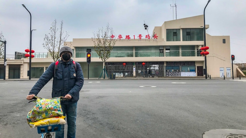 A man in a black face mask stands with a bicycle in front of an empty street and building with hanging red lanterns.