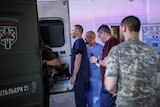 Three people in scrubs stand at the back of an ambulance looking inside 