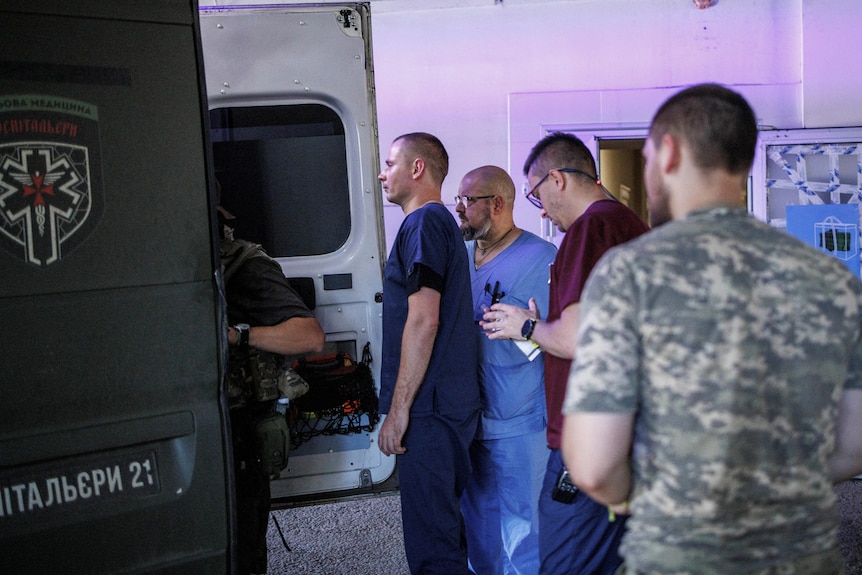 Three people in scrubs stand at the back of an ambulance looking inside 