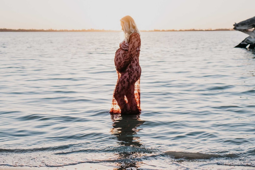 Woman with a pregnant belly stands in shallow water of lake with sun setting behind her.