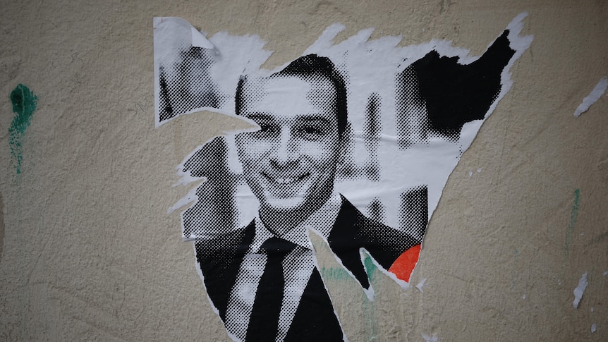 A poster with a picture of a young man smiling is partially torn of a wall 