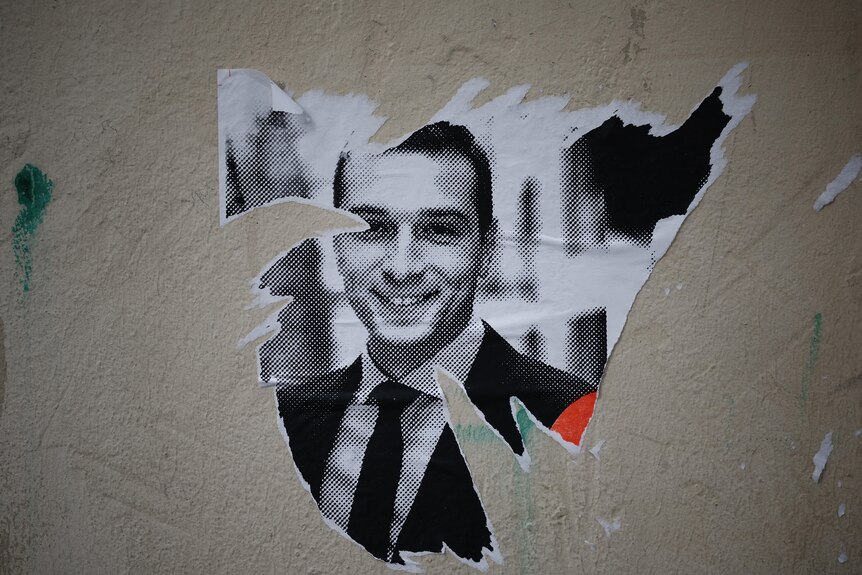 A poster with a picture of a young man smiling is partially torn of a wall 