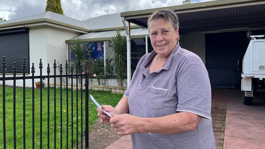 A woman wearing a purple polo shirt standing in front a house while holding a letter