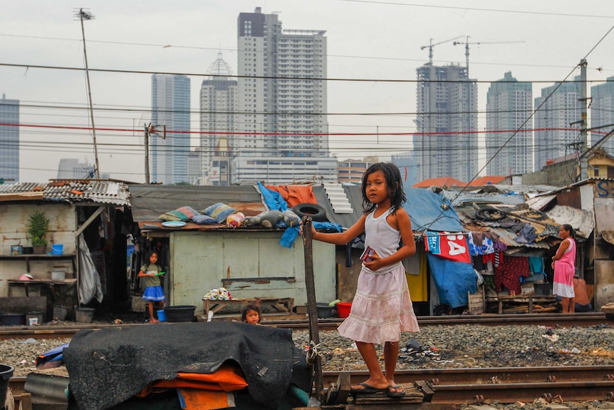 A little girl stands on train tracks near slums with skyscrapers in the background