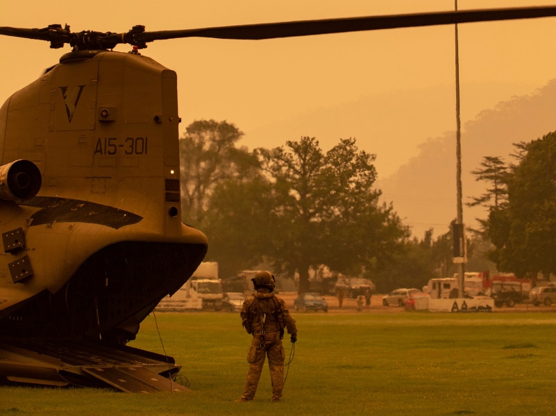 An Australian Army soldier watches a Blackhawk helicopter leave Omeo showgrounds surrounded by a yellow sky.