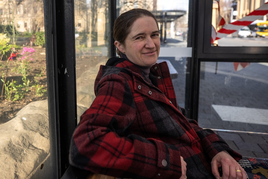 A woman with dark hair and a red and black checked jacket sits in a glass bus shelter in front of a garden bed.