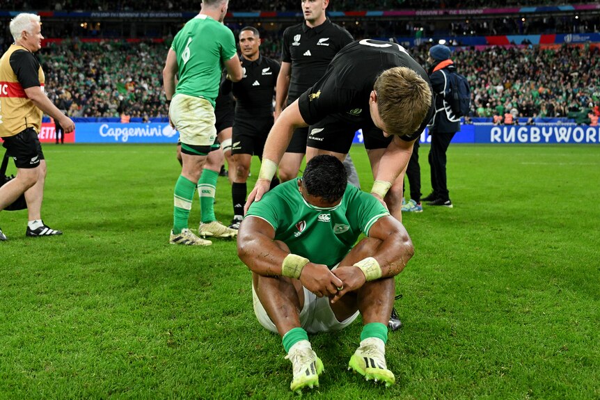 Jordie Barrett leans over to help Bundee Aki, who is sitting on the ground with his jersey over his face