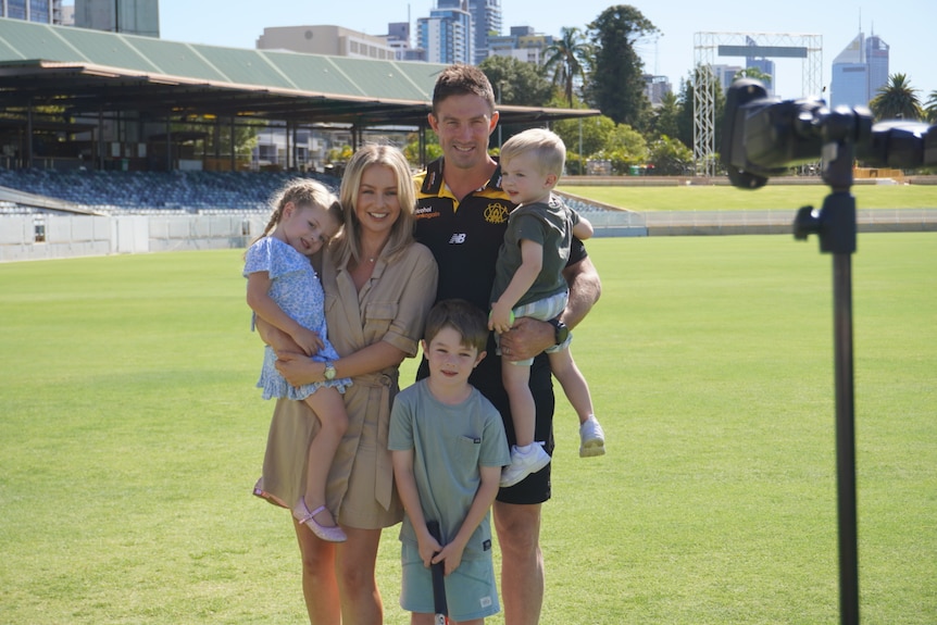 A family with a man and wife and three children standing on a large grassed over with a grandstand in the background