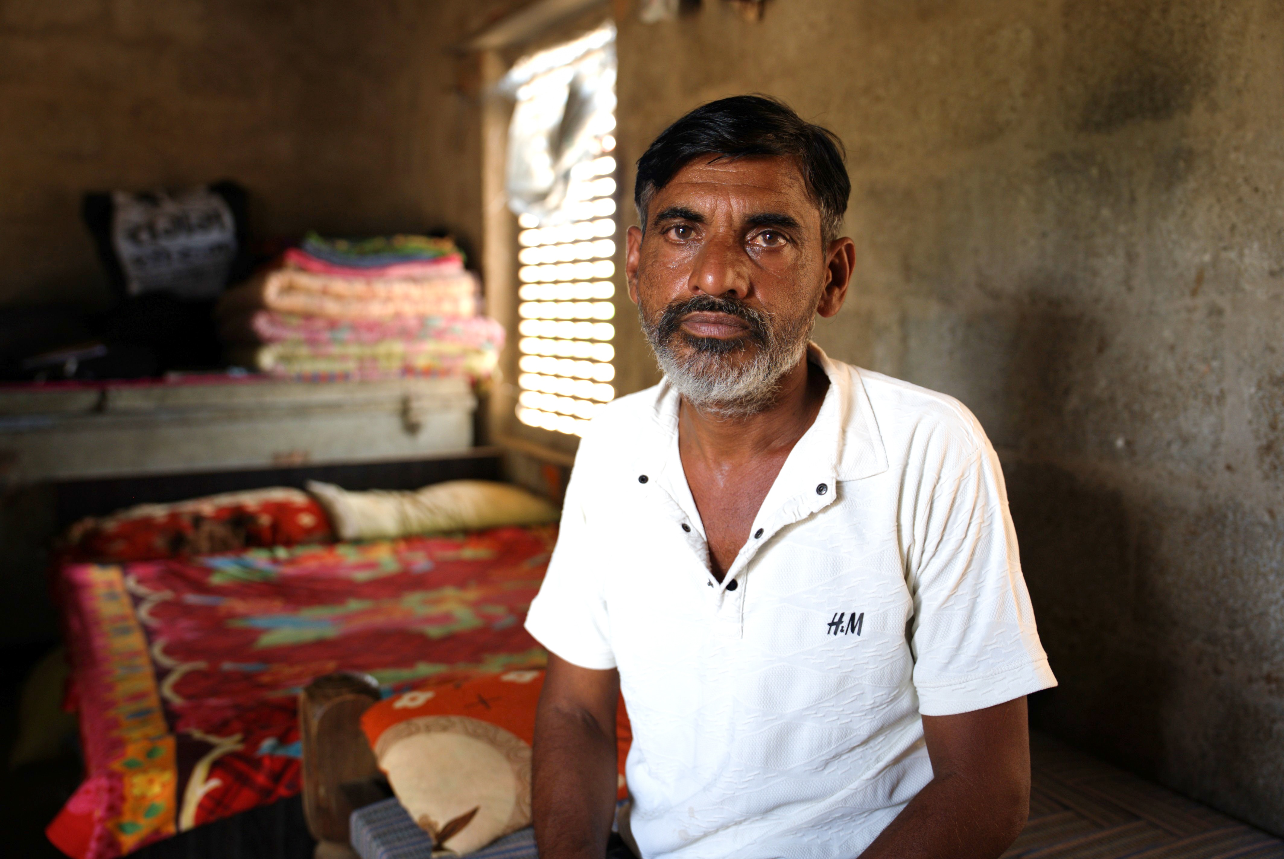 A man sits on a bed in a modest home, with a solemn expression.