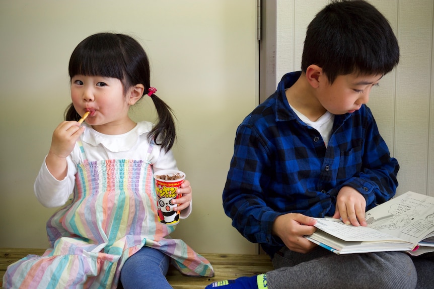 A little Japanese girl eats a snack while her brother reads a book