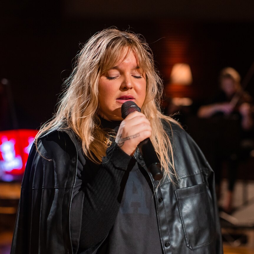 Tones is in a black jacket with long, blonder hair. She is singing into a microphone.