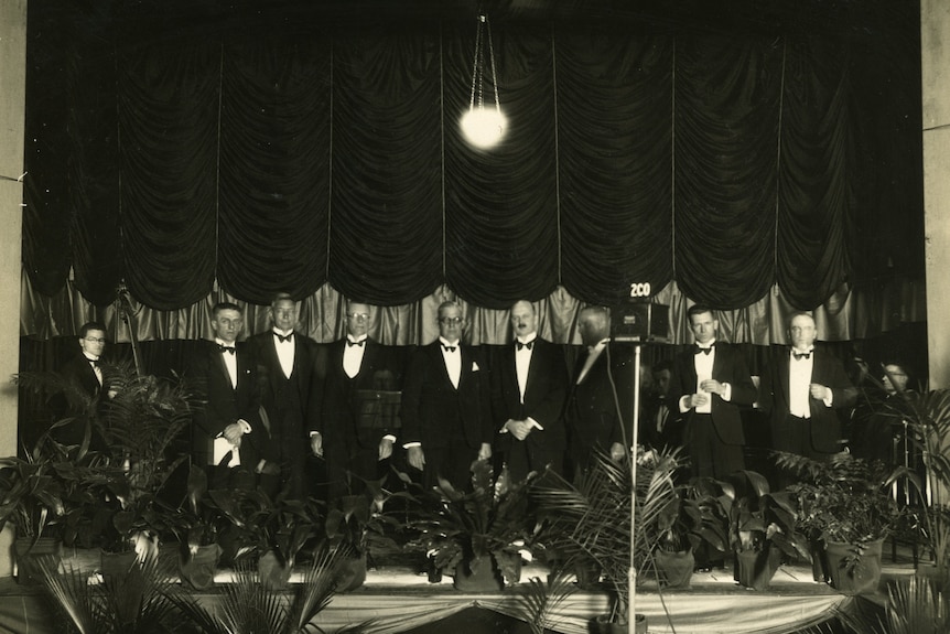A black and white photo of men in tuxedos in a fancy hall.