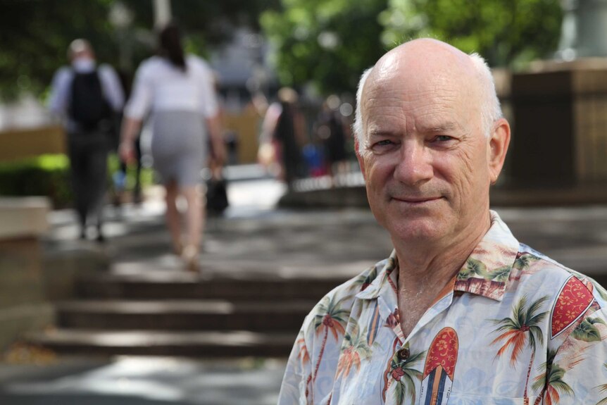 A bald, white haired man wearing a Hawaiian shirt looks at the camera, smiling, while standing outside on a sunny day.