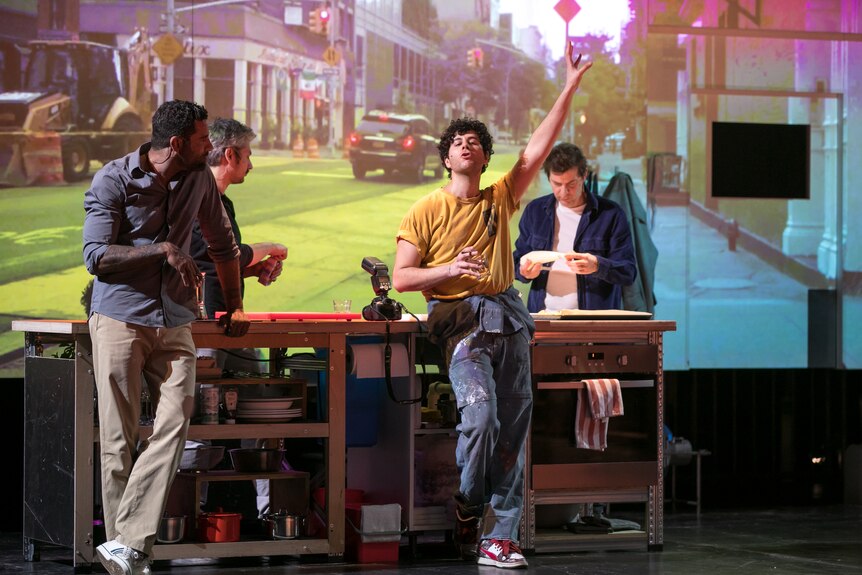Four male actors are gathered around a kitchen island bench on a stage with a street projected onto the wall behind them.