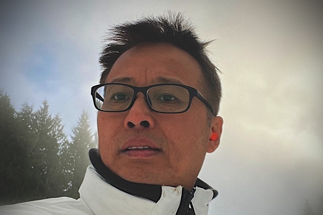 A man in glasses and a warm jacket takes a selfie with trees in the background