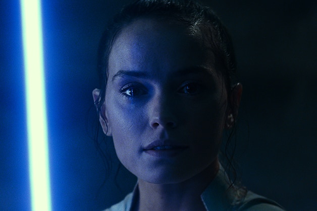 A woman stands in the dark, her face illuminated from one side by bright blue lightsaber.