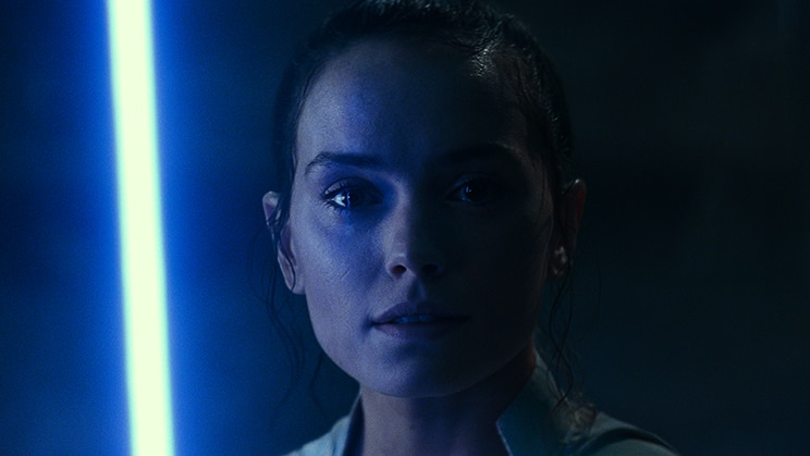 A woman stands in the dark, her face illuminated from one side by a bright blue lightsaber.