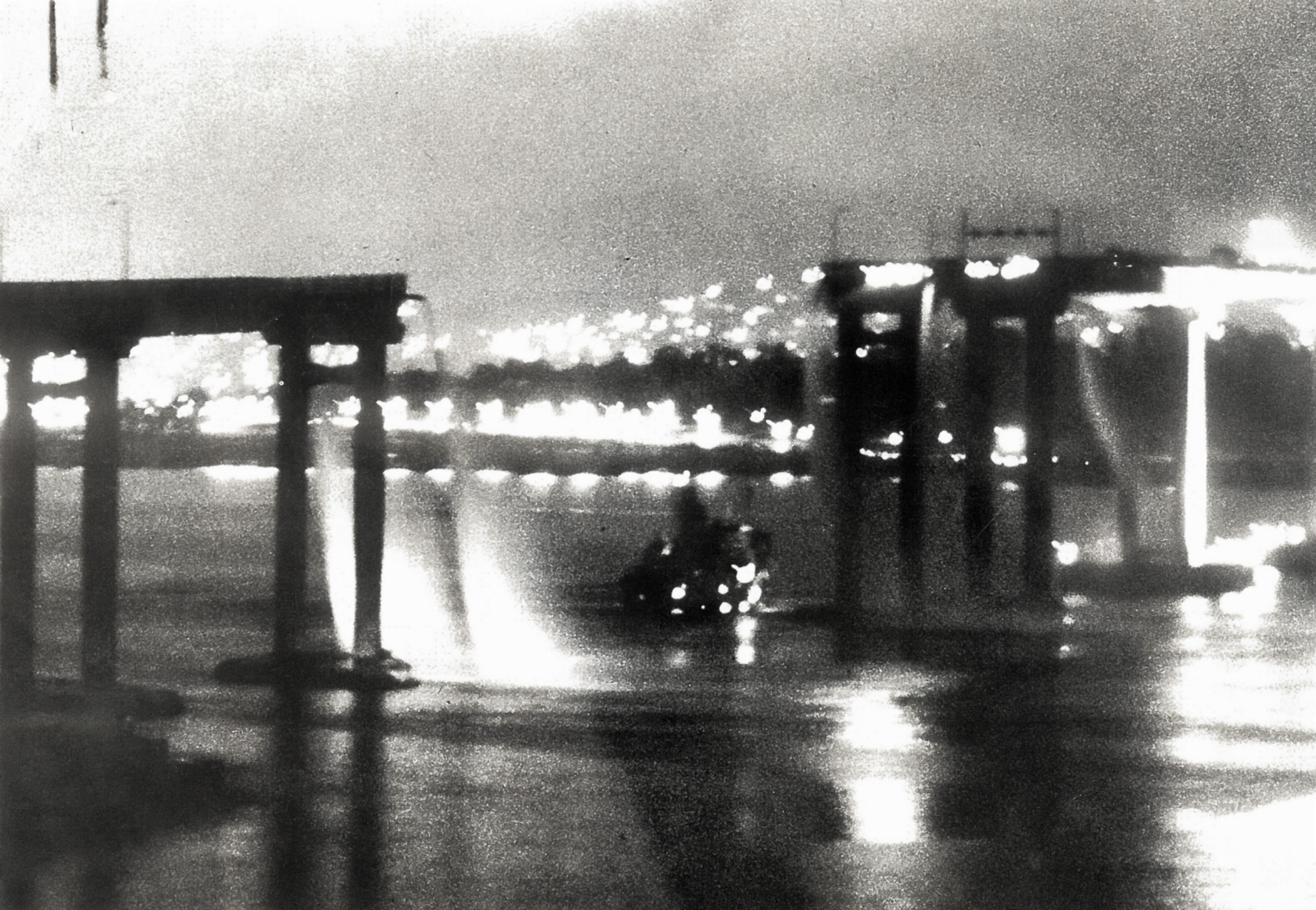 A black and white blurry photo at night of the top of a ship barely visible above the water next to a destroyed bridge.